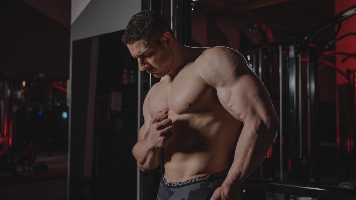 A muscular man flexing his physique under dramatic lighting in an indoor gym setting, showcasing fitness and strength.